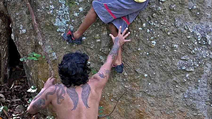 A father is keeping a close watch over his climbing son on an outdoor boulder. He stands braced, ready to assist his child if needed.