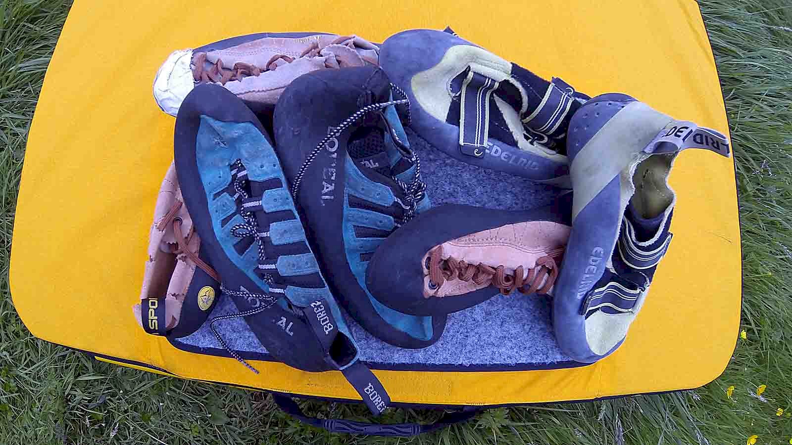 The picture shows a pile of climbing shoes of varying ages spread out on a bouldering pad.