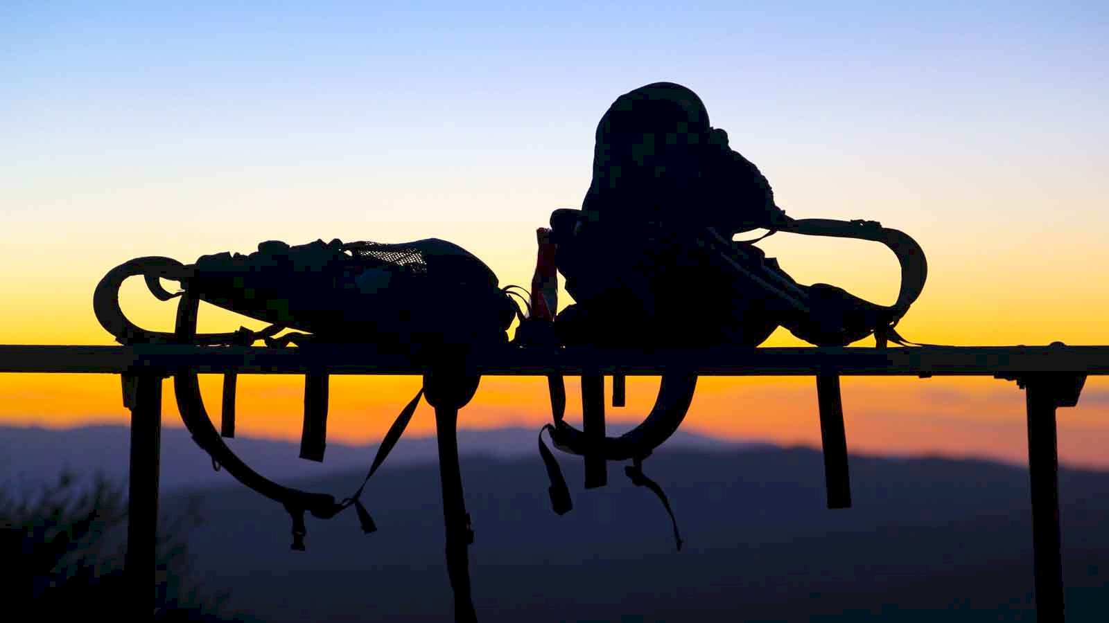 Picture of two backpacks balancing on a railing in the foreground with a orange sunset sky illuminating the hills in the distance.
