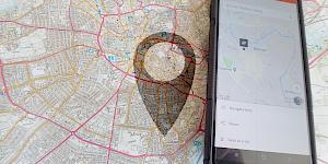 Image showing a 1:25000 OS map with a phone showing What 3 Words beside it, faded with a location symbol.