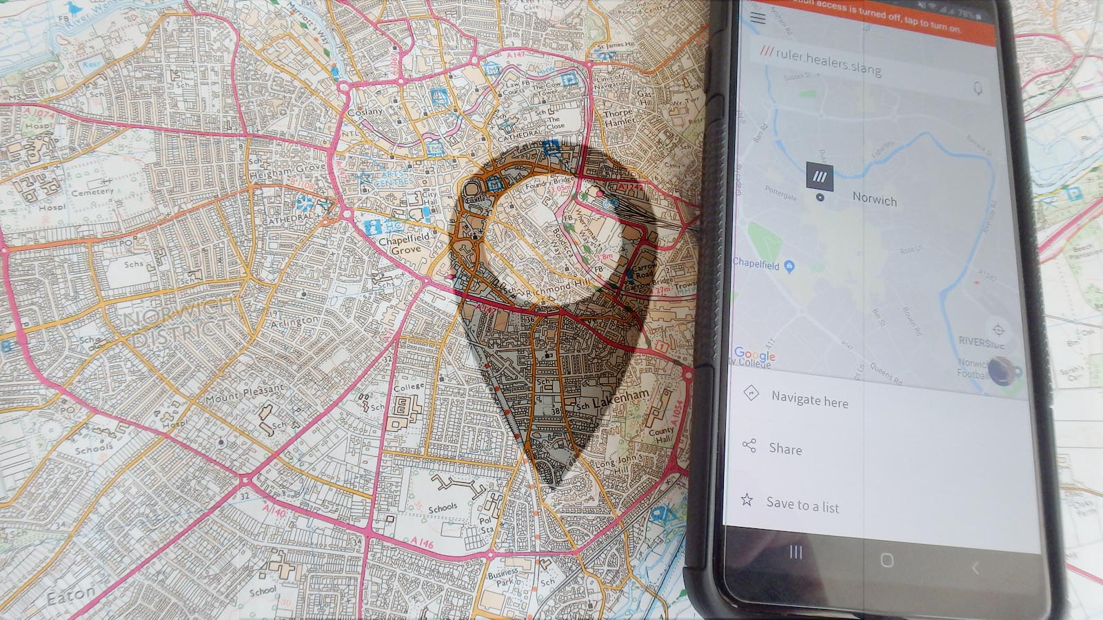 Image showing a 1:25000 OS map with a phone showing What 3 Words beside it, faded with a location symbol.